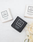Three square soap boxes branded with Só, one white labelled Honey Oat Cleansing Bar, one Black labelled Charcoal Cleansing Bar and another white one labelled Lather Gentle Cleansing Bar styled on a marble background with faux florals.