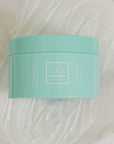 A mint green plastic jar of Só Butter Coco Mint body butter, placed on top of a textured background of white whipped product