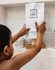 A child in a bathroom stands in front of a built-in shelf, holding a white pouch of Só Coconut & Oak Bath. The scene includes a white tiled wall, a glass jar of bath salts, and another Só product container on the shelf.