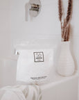 White bag with 'Só Flaky' branding on the edge of a bathtub with a cream coloured vase with dried florals.