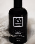 A black bottle of Só Soap hand wash concentrate in Citrus Pepper scent, standing upright styled on a blurry white background with pampas grass and cloth.