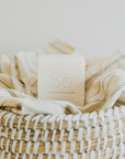 A square white bar of soap with the brand name 'Só' embossed on top, styled on a straw basket with a linen coloured cloth