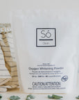 A package of Só Clean Oxygen Whitening Powder with a small scoop and a cloth beside it, on a white background with eucalyptus leaves.