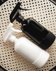 Two spray bottles, one black with a silicone sleeve and one white with a silicone sleeve, on a woven textured surface.