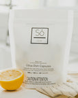 A white package of Só Clean Citrus Dish Capsules with a half lemon beside it, indicating a citrus scent on a blurred light background with a beige fabric.