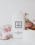 A white plastic deodorant style container branded Só Salve Ice Salve a cooling muscle salve in 50 g twist-up stick styled on a white backdrop with the lid off showing a white product and natural stone and floral elements.