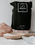 A package of Só Salty bath soak with Epsom salt and Himalayan pink salt, labelled Coco Mint with a wooden spoon with pink salt crystals and natural clear and pink stones.