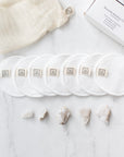 White reusable cotton face pads with small sewn-on 'Só' branded tags, arranged in a row with a cream mesh bag next to a white box on a light marble surface with a few crystals.