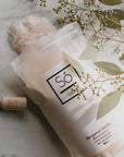 A package of Só Salty bath soak with Epsom salt and Himalayan pink salt labelled Bergamot Vanilla styled with a wooden scoop, pink salt cystals spilling out of the bag and eucalyptus branches.