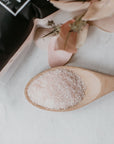 A package of Só Salty (Essential Oil) bath soak with Epsom salt and Himalayan Pink salt, scented with various essential oils, displayed with a small amount of pink salt visible in a wooden spoon surrounded by decorative florals.
