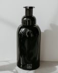 A black glass foaming soap dispenser with a pump, placed on a marble surface and white background with a shadow falling over it.