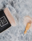 A clear bag of pink salt labelled Só Salty epsom and himalayan pink salt bath soak styled with a wooden scoop and some pink salt crystals on a grey marble backdrop