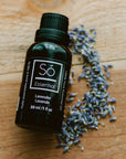 A black bottle of Só Essential Oil labelled Lavender 30 ml on a wood surface with lavender buds