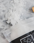 A bag of Só brand magnesium bath flakes spilled open with the contents, a white crystalline substance, scattered on a grey marble surface with a wooden Só branded scoop.