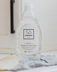 An oval natural loofah sponge with a bottle of Só Clean Tangerine Dish Soap styled on a kitchen counter with a black cupboard handle in the background and a blue and white striped tea towel.