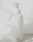 A white, glass foaming soap dispenser with a pump, placed on a marble surface and white background with a shadow falling over it.