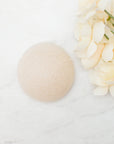 A cream coloured round konjac sponge, on a white marble surface next to cream-colored flowers.