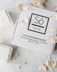 A bath bomb from Só Bomb with packaging that reads Coco Oat Bath Bombs' The product is displayed on a white marble backdrop with light pink flower petals and completed bath bombs with a powdered substance sprinkled on both the completed bath bombs and the branded box.