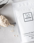 A package of Só Luxury Coco Oat Milk Bath powder with some scattered on a gold spoon beside it, all placed on a white marble surface.