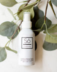A white spray bottle labeled 'Só Outdoorsy' placed against a backdrop of green leaves, representing an outdoor body spray product.