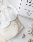 White reusable cotton face pads with small sewn-on 'Só' branded tags, arranged on a cream mesh bag next to a white box on a light marble surface with a few crystals.