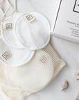 White reusable cotton face pads with small sewn-on 'Só' branded tags, arranged on a cream mesh bag next to a white box on a light marble surface with a few crystals.