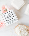 A styled flat-lay of a white box of Só Bomb Coco Oat Bath Bombs on a light marble surface, surrounded by pink roses, finished bath bombs, and a wooden spoon filled with the white powder.