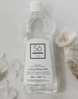 A bottle of Só Clean Citrus Rinse Aid on a marble surface styled with naturlal rocks and flowers.