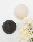 Two round konjac sponge, one cream coloured and one dark charcoal gray, arranged on a white marble surface next to cream-colored flowers.