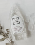 A clear bottle of Só Clean Tangerine Dish Soap with a white label, surrounded by some plant leaves and clear rocks on a white background.