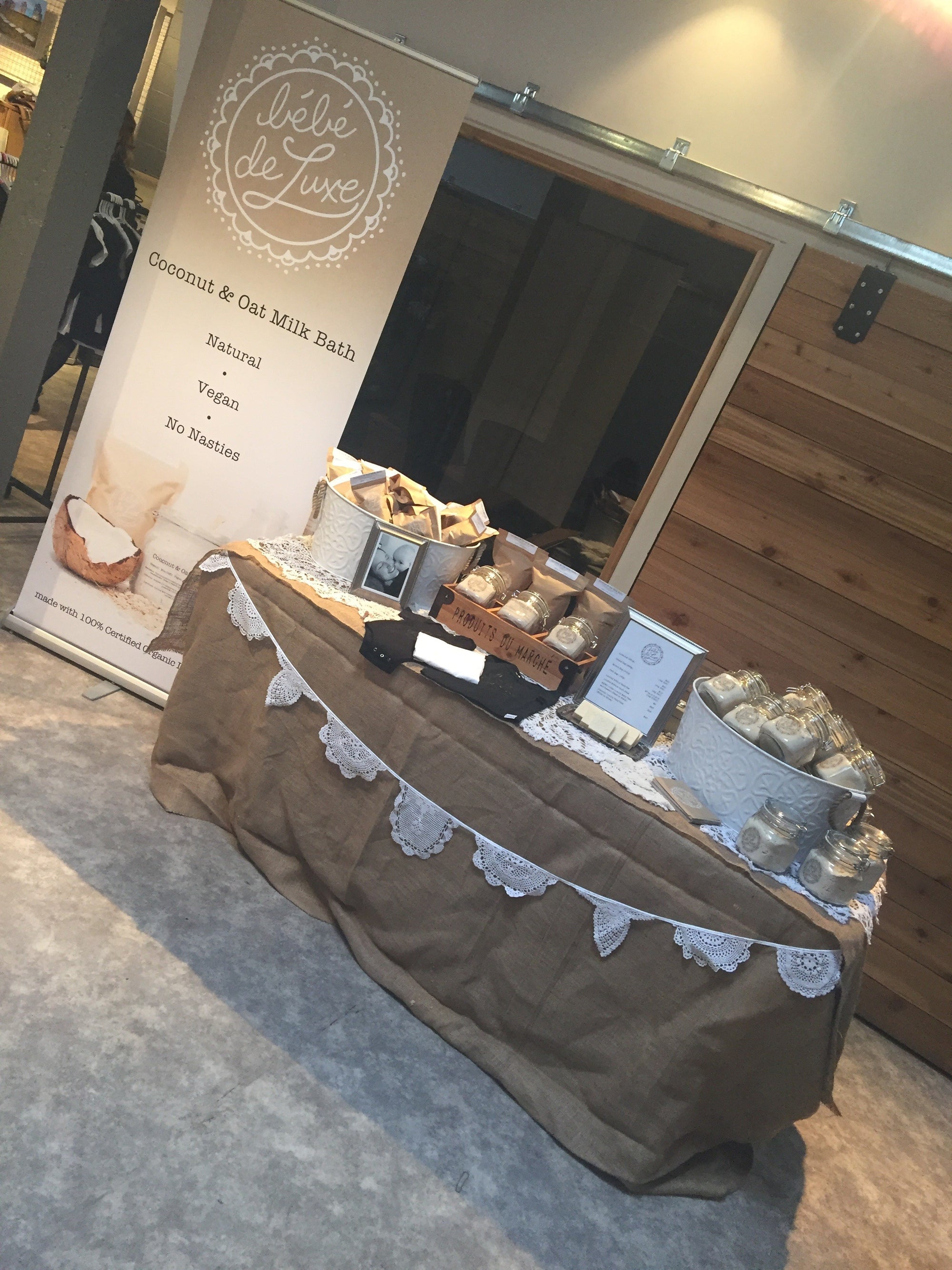 A product display table for 'Bebe de Luxe' Coconut & Oat Milk Bake mix, featuring a brown burlap tablecloth with a white lace bunting garland, alongside product containers and a promotional banner on a carpeted indoor floor.