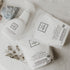 Three white Só Clean laundry and stain care products arranged on a light marble surface: a stain remover spray bottle, a powdered laundry soda bag, and a whitening powder bag, with decorative stones and dried flowers nearby.