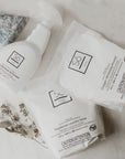 Three white Só Clean laundry and stain care products arranged on a light marble surface: a stain remover spray bottle, a powdered laundry soda bag, and a whitening powder bag, with decorative stones and dried flowers nearby.
