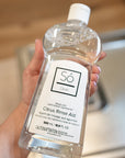 A hand holding a clear bottle with a clear liquid and white Só branded label listing the product as Citrus Rinse Aid, an open dishwasher can be seen in the blurred background.