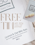 A close-up of a white product package for “FREE TIN” coconut and oat milk bath, with a small white tin shown beside it on a soft beige textured surface. The package text reads “Coconut & Oat Milk Bath,” “300 g,” and the visible brand is “Só