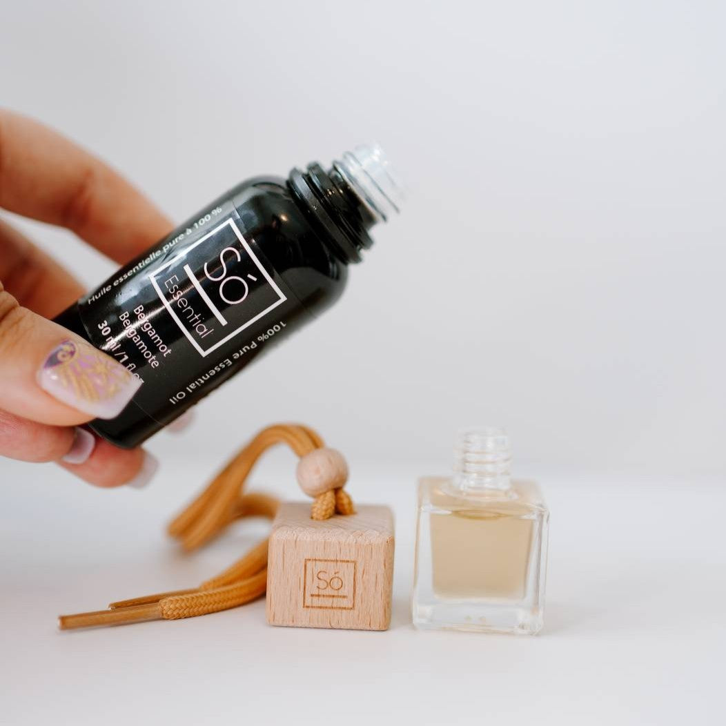 A hand holding a black bottle of Só branded essential oil to pour it into a small glass jar with the Só branded wooden lid off 