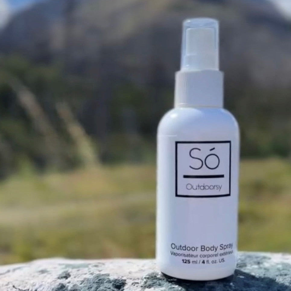 A white bottle of Só Outdoorbody spray standing on a gray stone surface with a blurred mountain landscape in the background.