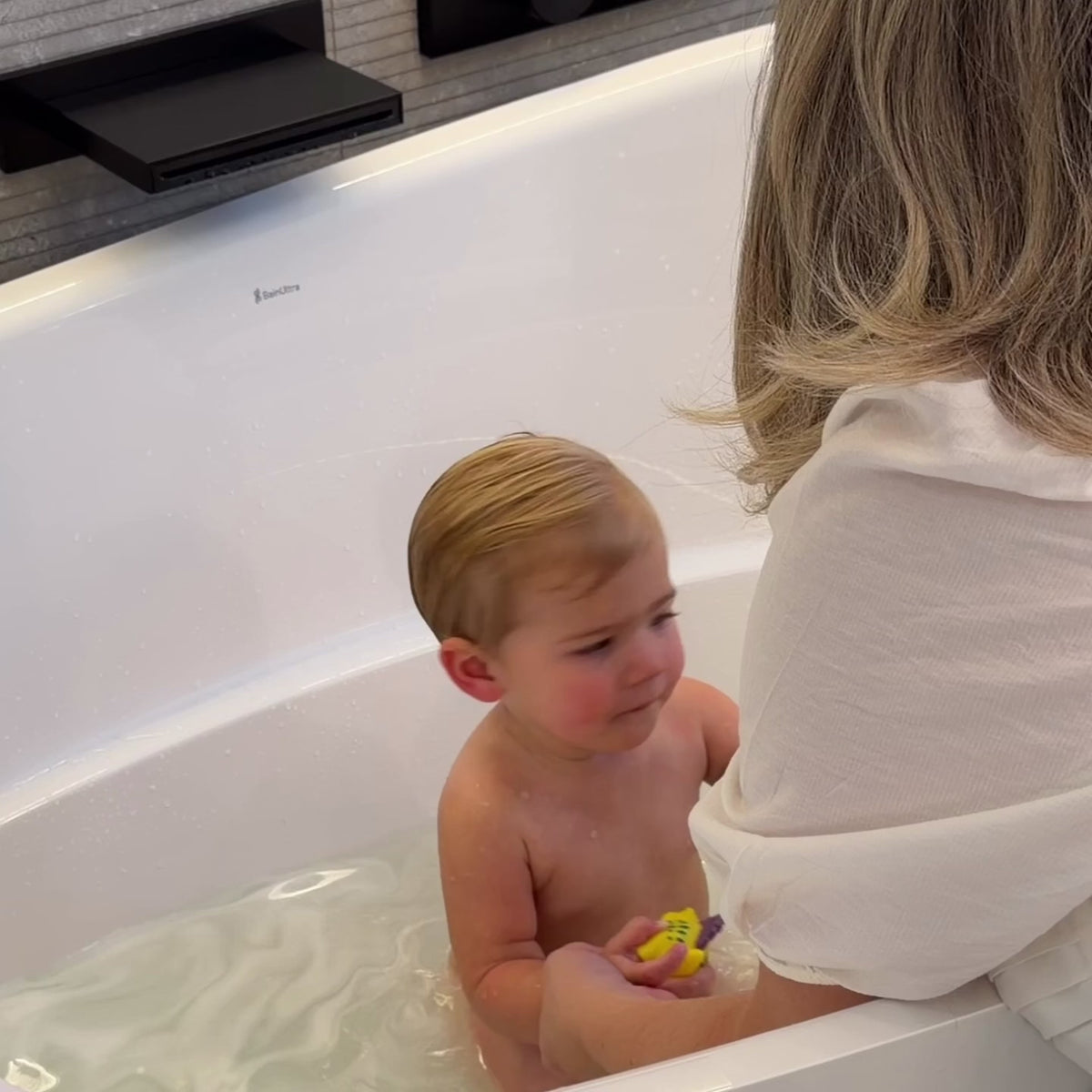 A mom in a white blouse with blonde hair bathing a blonde baby in a white bathtub using a konjac sponge to wash