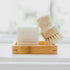 A bamboo soap dish holding a white bar of soap embossed with the brand 'Só' and a small wooden-handled cleaning brush with natural bristles, set on a bright white surface near a softly blurred window.