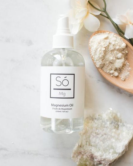 A clear spray bottle labeled 'Só  Magnesium Oil' against a marble background withnatural stone and floral elements with a wooden spoon with a beige powder.