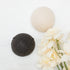 Two round konjac sponge, one cream coloured and one dark charcoal gray, arranged on a white marble surface next to cream-colored flowers.